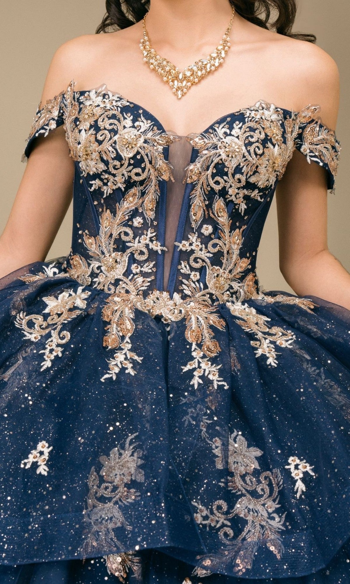 A woman wearing the Quinceanera Dress 8390JQ by Cinderella Couture—an off-the-shoulder navy ball gown adorned with gold floral embroidery, glitter accents, and paired with a gold statement necklace.